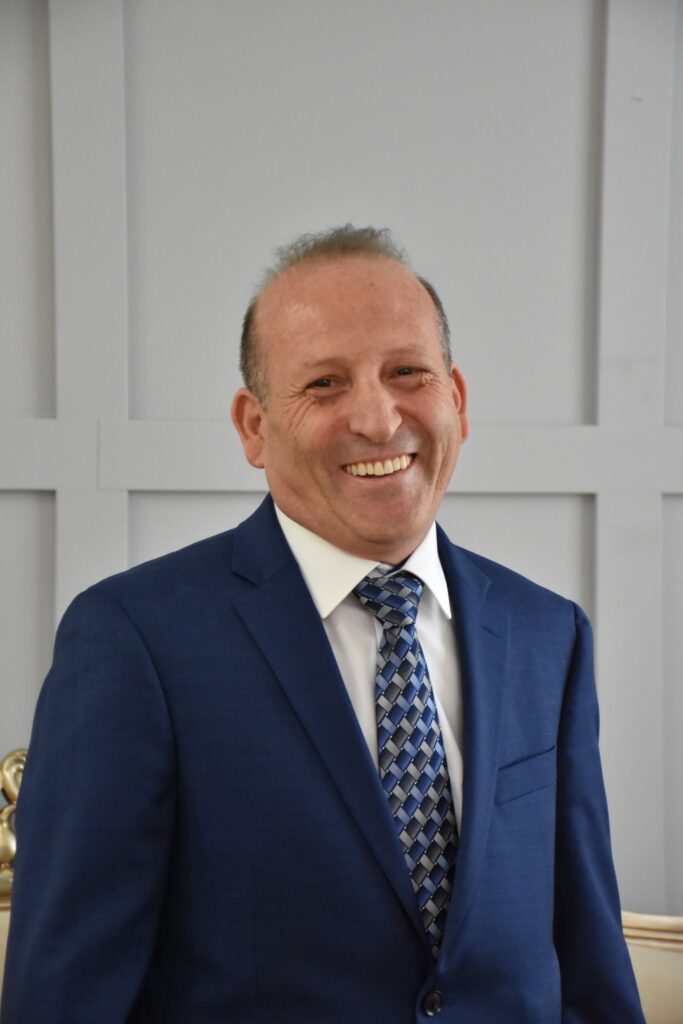 A smiling middle-aged man in a navy blue business suit with a patterned blue and gray tie, standing in what appears to be an office or professional setting with white paneled walls in the background.
