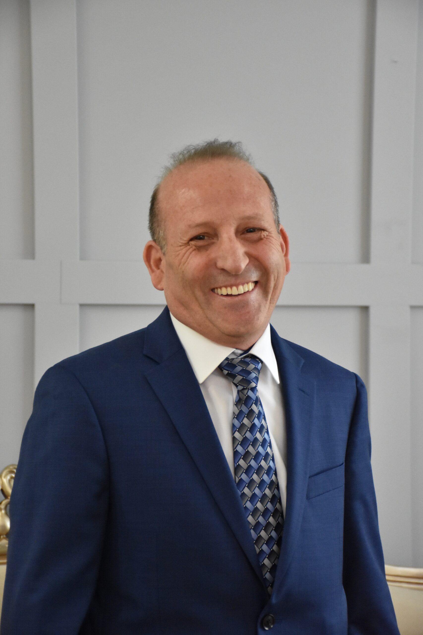 A smiling middle-aged man in a navy blue business suit with a patterned blue and gray tie, standing in what appears to be an office or professional setting with white paneled walls in the background.
