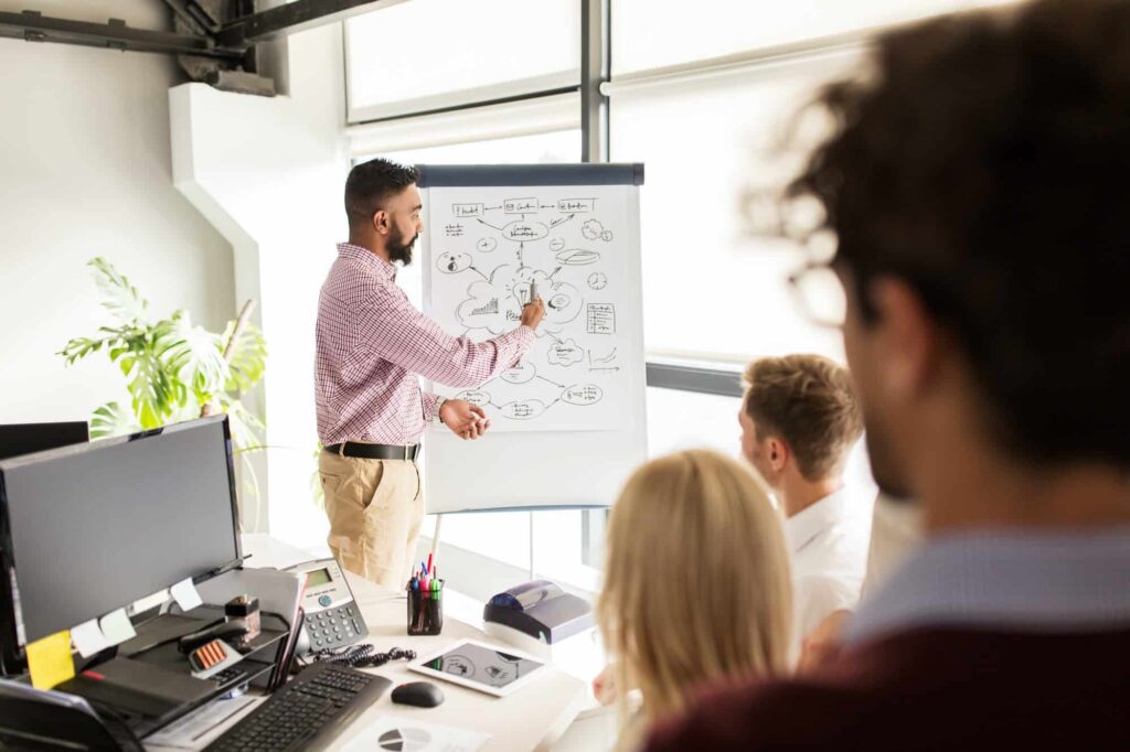 A man in a checkered shirt and khaki pants presenting at a whiteboard with diagrams and flowcharts to a small group of colleagues in a modern office setting. The presenter is pointing to the board while several people sit at a desk with computers, viewed from behind one of the attendees.